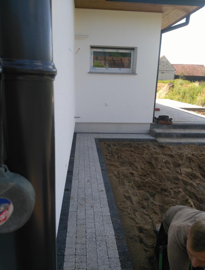 Fragment posesji w trakcie prac brukarskich: ułożony pas kostki granitowej obrzeżony ciemniejszym materiałem, obok teren przygotowany pod dalsze prace, widoczny pracownik w tle, rynna i budynek w tle.
