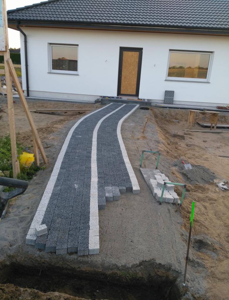 Ścieżka z kostki granitowej w trakcie układania, z białymi obrzeżami, prowadząca do drzwi wejściowych nowoczesnego domu jednorodzinnego, widoczny wykop ziemny obok.