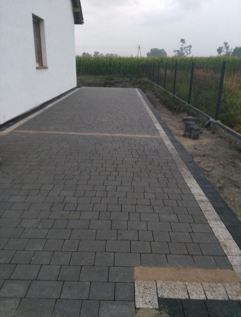 Wykończona nawierzchnia z ciemnej kostki brukowej przed białym budynkiem, z obrzeżem z jasnej kostki granitowej i widocznym fragmentem ogrodzenia w tle.