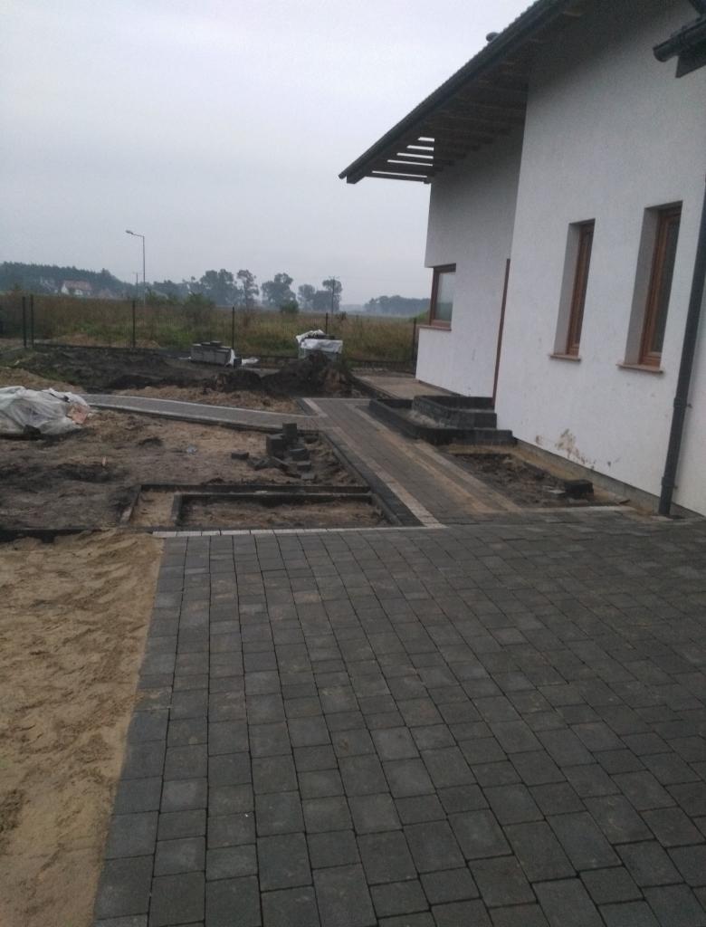 Częściowo ułożona, grafitowa kostka brukowa przed białym domem z brązowymi ramami okiennymi; widoczne przygotowane podłoże i piasek, w tle pole i pochmurne niebo.