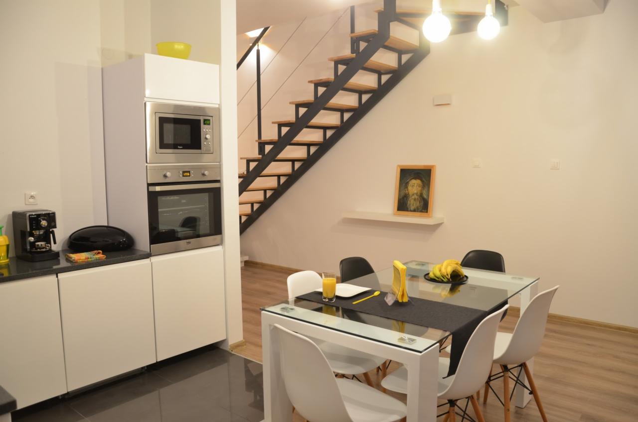 Jasne wnętrze: biała zabudowa kuchenna z piekarnikiem i mikrofalówką, szklany stół z białymi krzesłami, schody z drewnianymi stopniami i czarną metalową konstrukcją, obraz na ścianie.