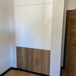 ANNA KUCNER - Biała szafa wnękowa z drewnianymi, pionowo żłobionymi frontami szuflad w dolnej części, czarne listwy przypodłogowe, drewniana podłoga i częściowo widoczne drewniane drzwi po prawej stronie.