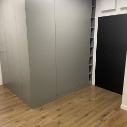 ANNA KUCNER - Szara szafa wnękowa z otwartymi półkami na boku, dopasowana kolorystycznie do ścian, na podłodze płytki imitujące drewno, obok czarne drzwi.