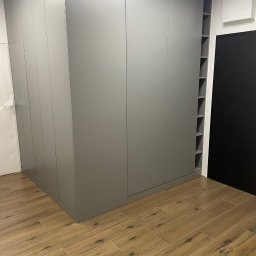 ANNA KUCNER - Duża, szara szafa wnękowa z otwartymi półkami z boku, dopasowana do podłogi z płytek imitujących drewno.