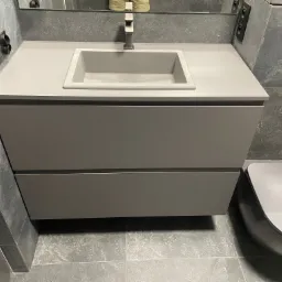 Szara, minimalistyczna łazienka z umywalką nablatową z szarego kamienia i szafką pod umywalką z dwiema szufladami, obok toaleta w tym samym odcieniu szarości i płytki imitujące kamień na ścianach...