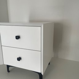 ANNA KUCNER - Biała szafka nocna z dwiema szufladami i czarnymi, kwadratowymi uchwytami, osadzona na cienkich, czarnych nóżkach, na tle białej ściany i podłogi.