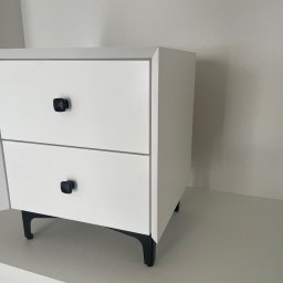 ANNA KUCNER - Biała szafka nocna z dwiema szufladami i czarnymi, kwadratowymi uchwytami, ustawiona na czarnych nóżkach w kształcie litery L, widoczna w rogu pokoju.