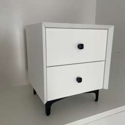 ANNA KUCNER - Biała szafka nocna z dwiema szufladami i czarnymi, kwadratowymi uchwytami, umieszczona na czarnych, skośnych nóżkach w minimalistycznym wnętrzu.