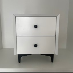 ANNA KUCNER - Biała szafka nocna z dwiema szufladami i czarnymi, geometrycznymi uchwytami oraz czarnymi nóżkami, ustawiona na białej półce w minimalistycznym wnętrzu.