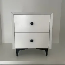 Biała szafka nocna z dwiema szufladami i czarnymi, geometrycznymi uchwytami oraz czarnymi nóżkami, ustawiona na białej półce w minimalistycznym wnętrzu.