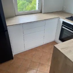 Nowoczesna, biała zabudowa kuchenna pod oknem z blatem w kolorze jasnego drewna, widok na zewnątrz przez okno, brązowa podłoga z płytek.