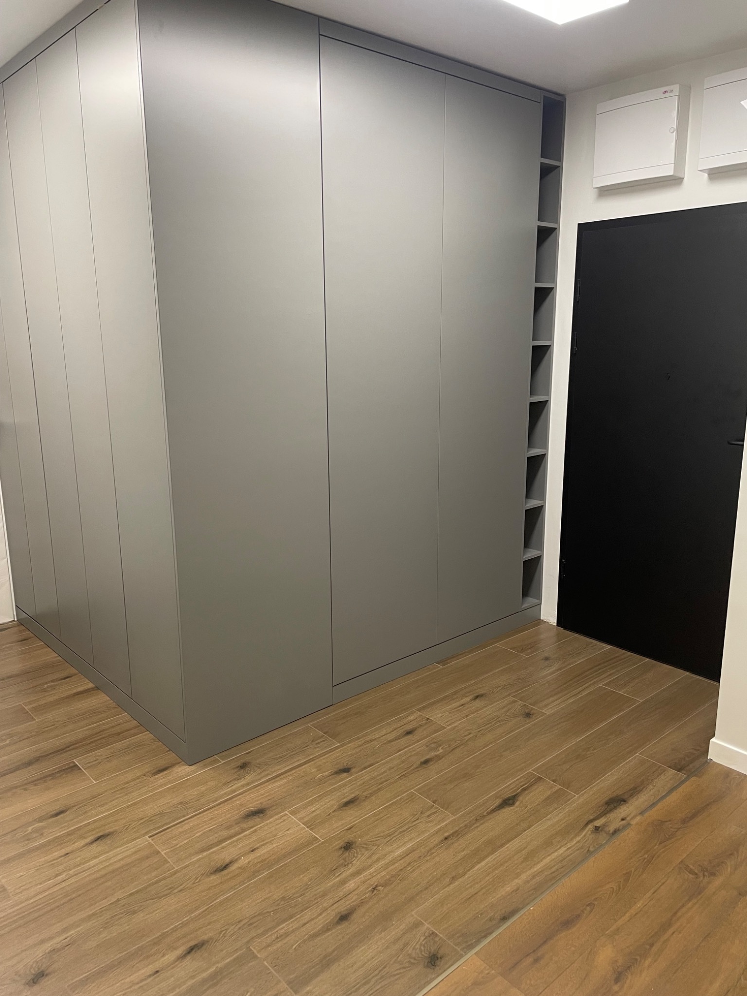Szara szafa wnękowa z otwartymi półkami na boku, dopasowana kolorystycznie do ścian, na podłodze płytki imitujące drewno, obok czarne drzwi.