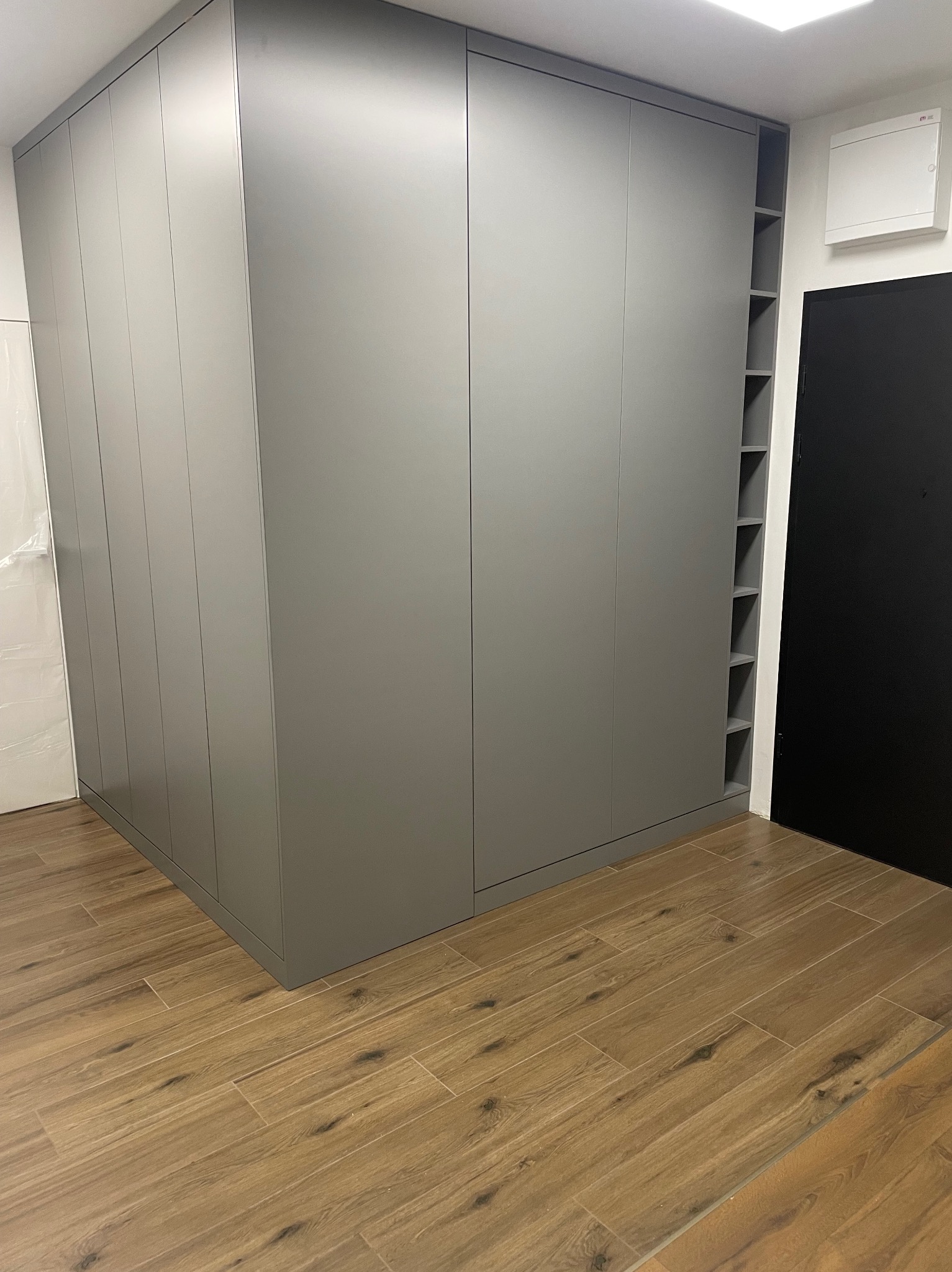 Duża, szara szafa wnękowa z otwartymi półkami z boku, dopasowana do podłogi z płytek imitujących drewno.