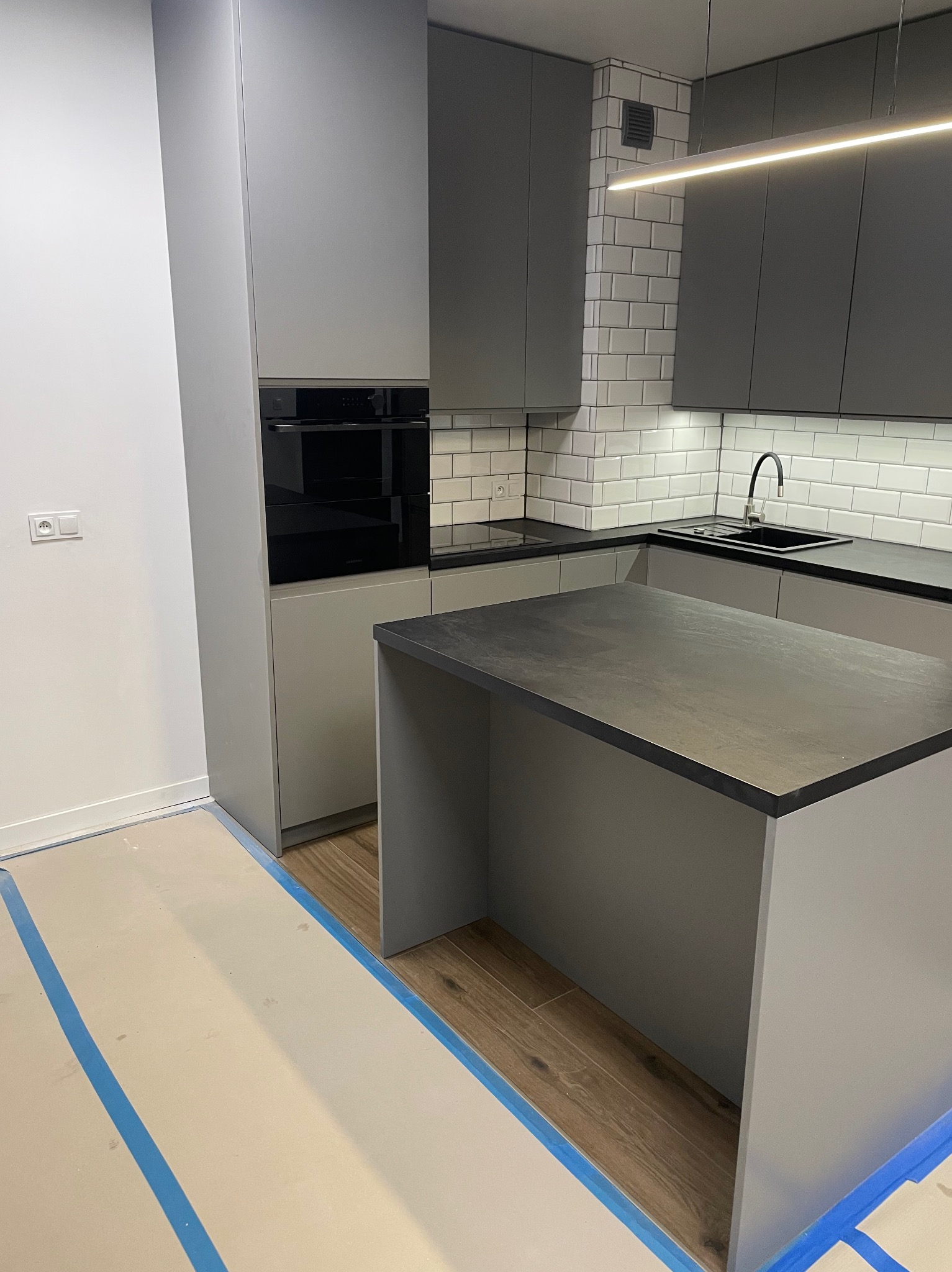 Nowoczesna, szara kuchnia z wyspą i białymi płytkami metra na ścianie, widoczne zabezpieczenia podłogi taśmą malarską, sugerujące prace wykończeniowe.