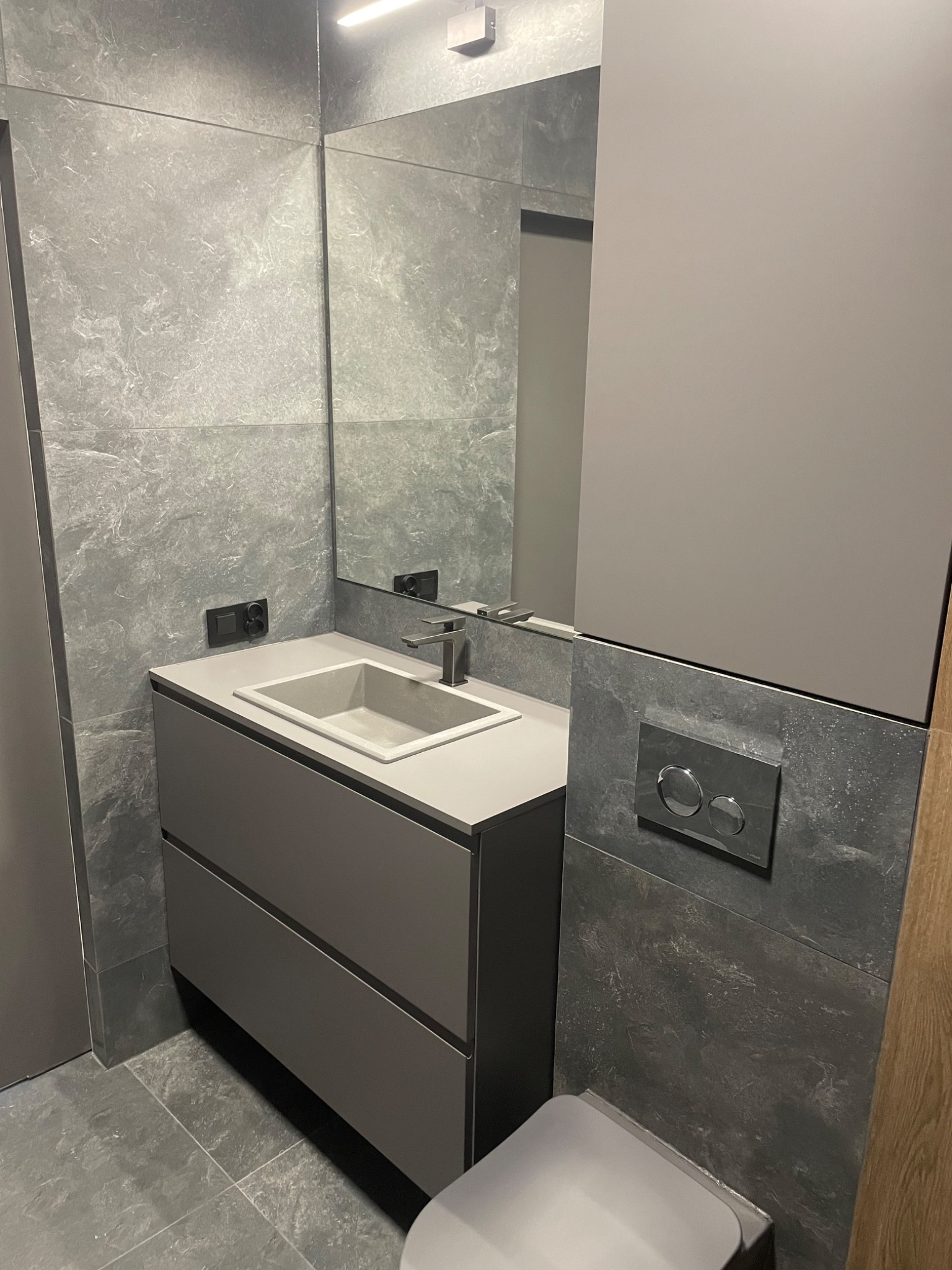 Nowoczesna łazienka z szarą zabudową umywalki z dwiema szufladami, umywalką z betonu architektonicznego, lustrem, szarą armaturą i płytkami imitującymi kamień.
