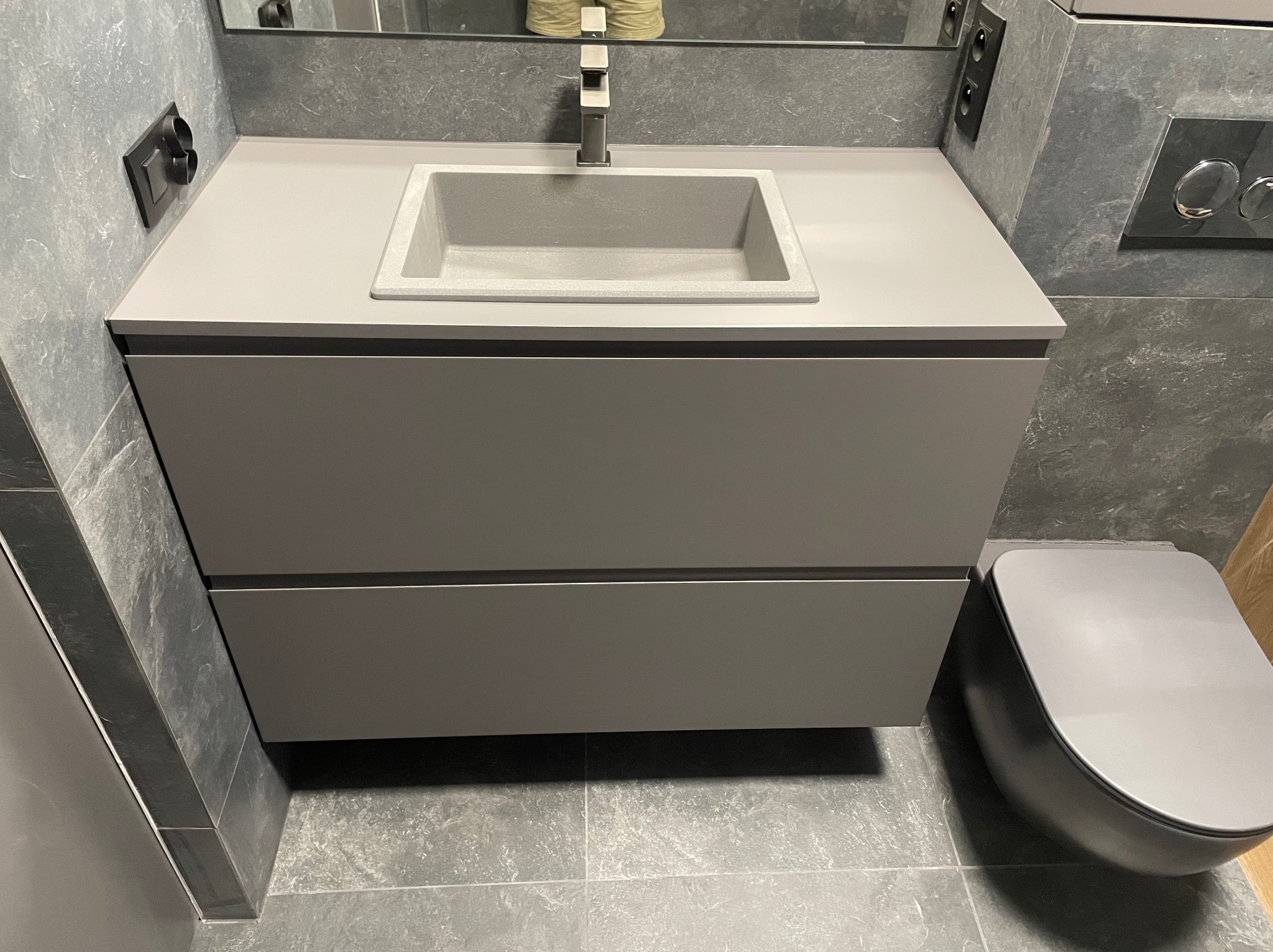 Szara, minimalistyczna łazienka z umywalką nablatową z szarego kamienia i szafką pod umywalką z dwiema szufladami, obok toaleta w tym samym odcieniu szarości i płytki imitujące kamień na ścianach...