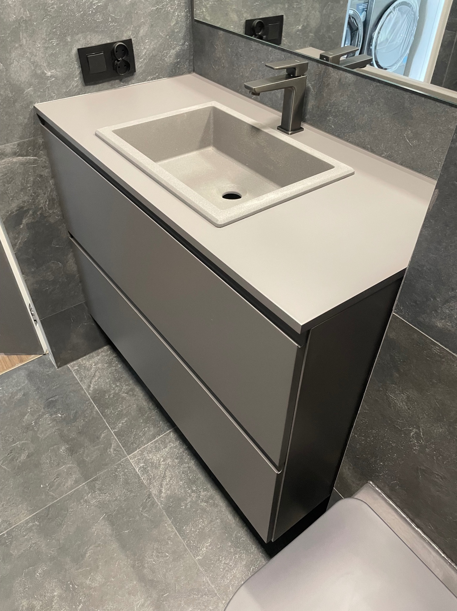 Szara szafka łazienkowa z umywalką i baterią w nowoczesnej łazience w Bujniczkach-Kolonii, widoczny fragment pralki w lustrze.