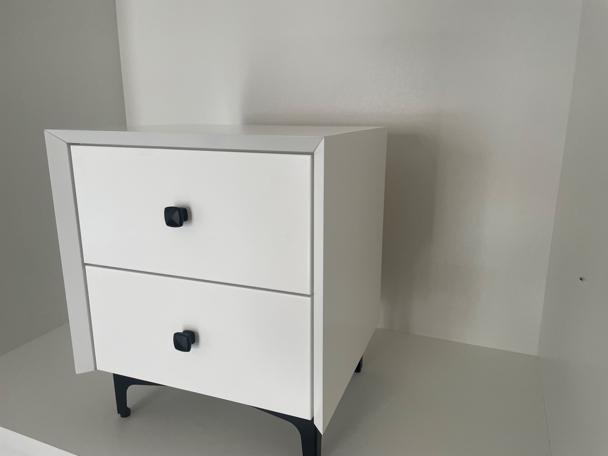 Biała szafka nocna z dwiema szufladami i czarnymi, kwadratowymi uchwytami, osadzona na cienkich, czarnych nóżkach, na tle białej ściany i podłogi.
