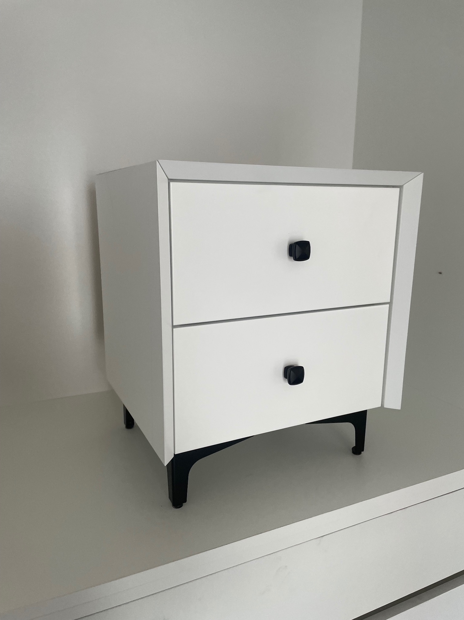 Biała szafka nocna z dwiema szufladami i czarnymi, kwadratowymi uchwytami, umieszczona na czarnych, skośnych nóżkach w minimalistycznym wnętrzu.