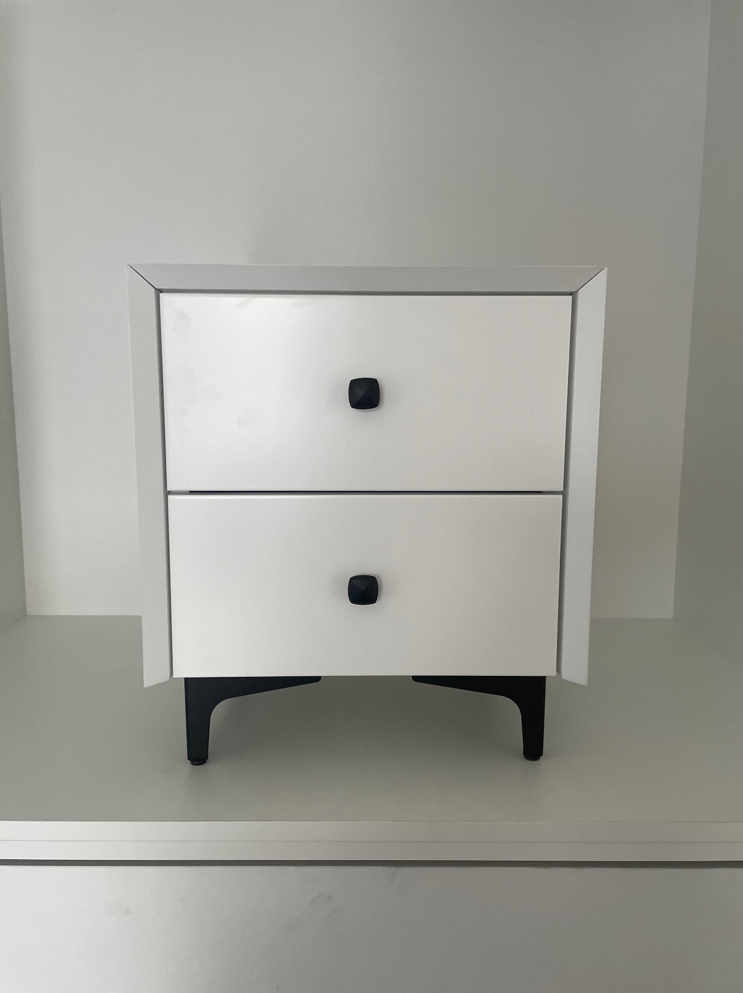 Biała szafka nocna z dwiema szufladami i czarnymi, geometrycznymi uchwytami oraz czarnymi nóżkami, ustawiona na białej półce w minimalistycznym wnętrzu.
