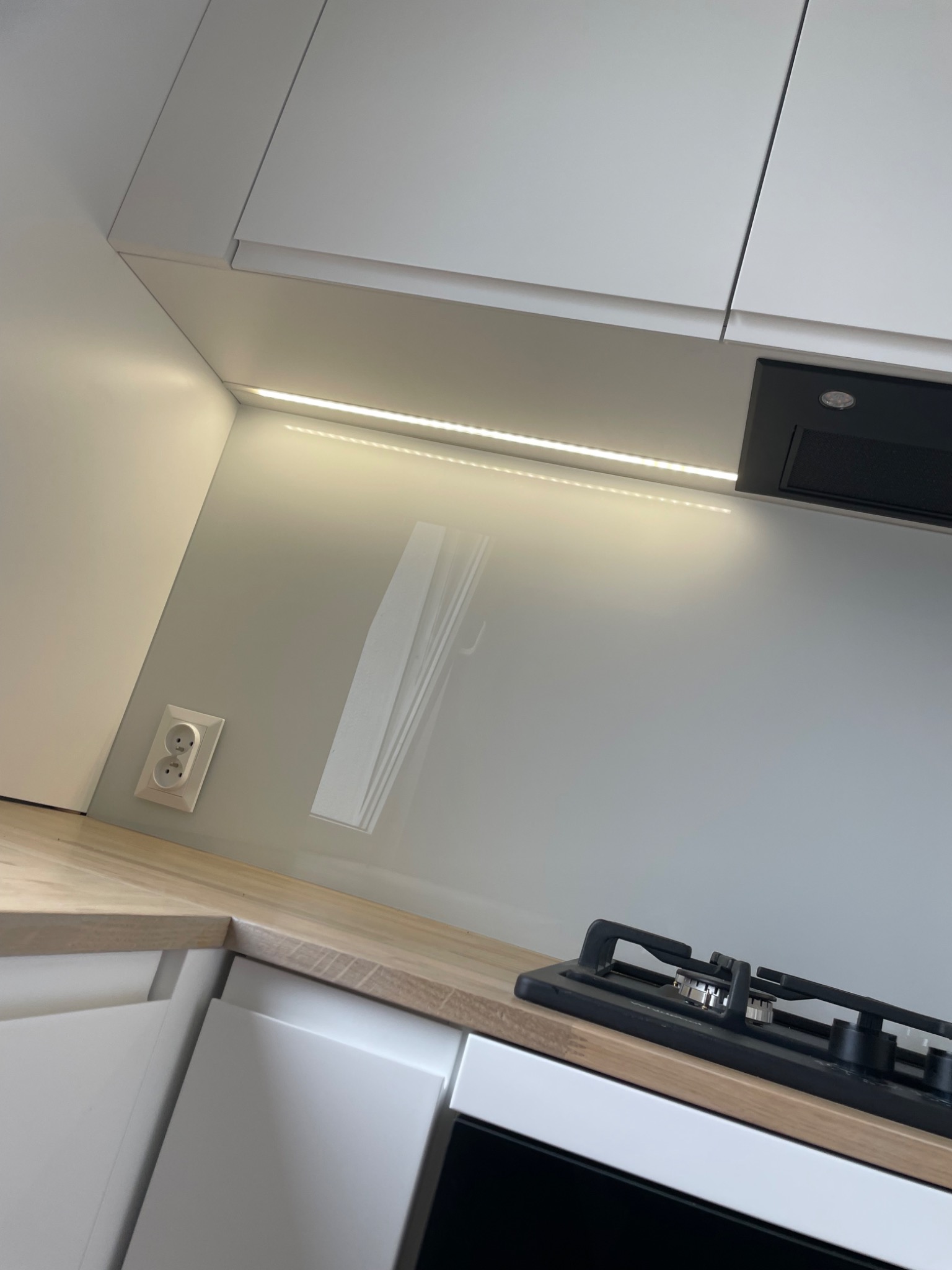 Fragment nowoczesnej kuchni z białymi szafkami, drewnianym blatem, szklanym panelem ściennym, oświetleniem LED podszafkowym i czarną płytą gazową.