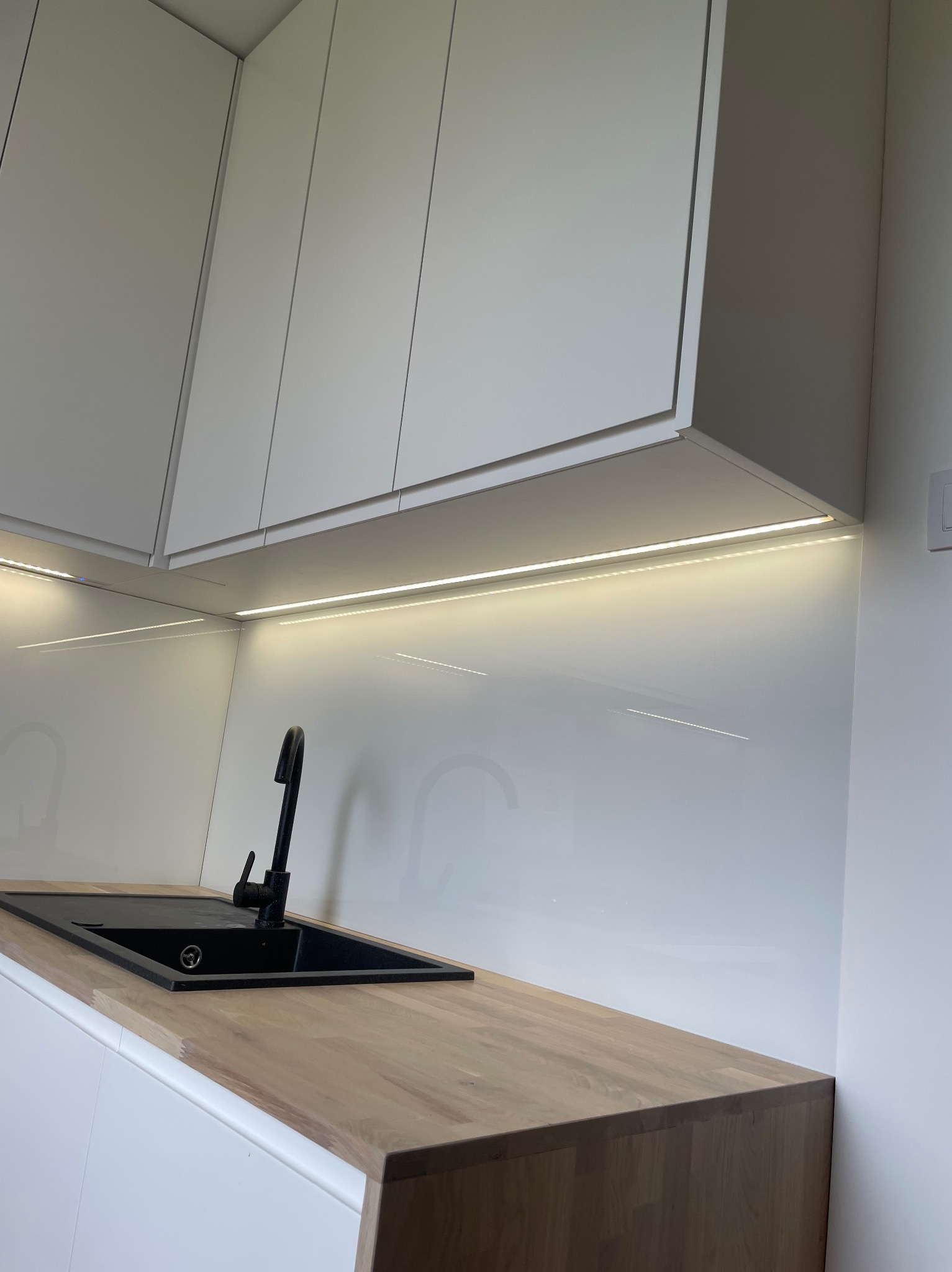 Nowoczesna kuchnia z białymi szafkami, drewnianym blatem i czarnym zlewem, oświetlona listwą LED pod szafkami.