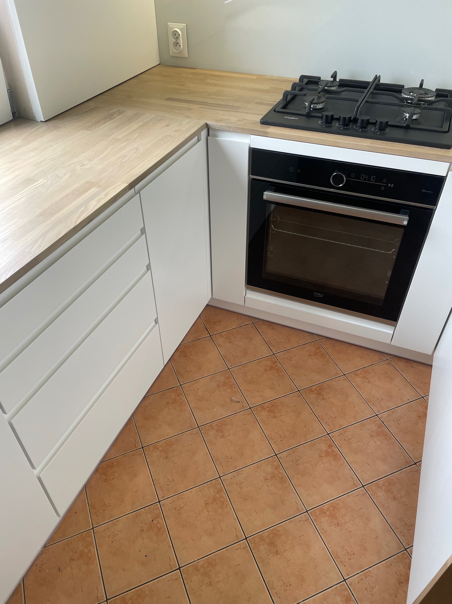 Fragment nowoczesnej, minimalistycznej kuchni z białymi frontami szafek, drewnianym blatem, wbudowanym piekarnikiem i płytą gazową na tle brązowej podłogi z płytek.