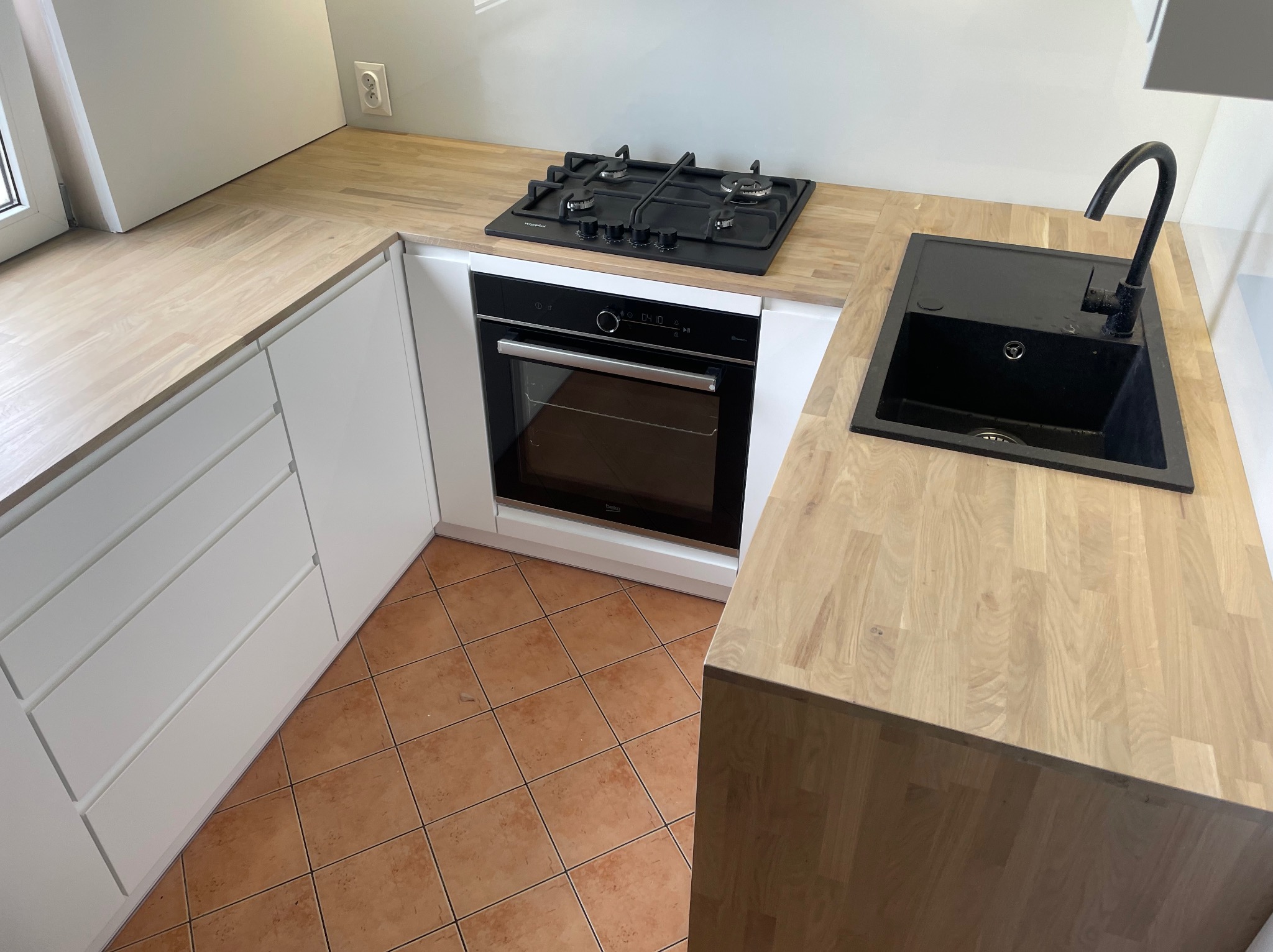 Nowoczesna kuchnia z jasnym drewnianym blatem, czarnym zlewem i płytą gazową oraz białymi szafkami z frontami bezuchwytowymi, pomarańczowa terakota na podłodze.