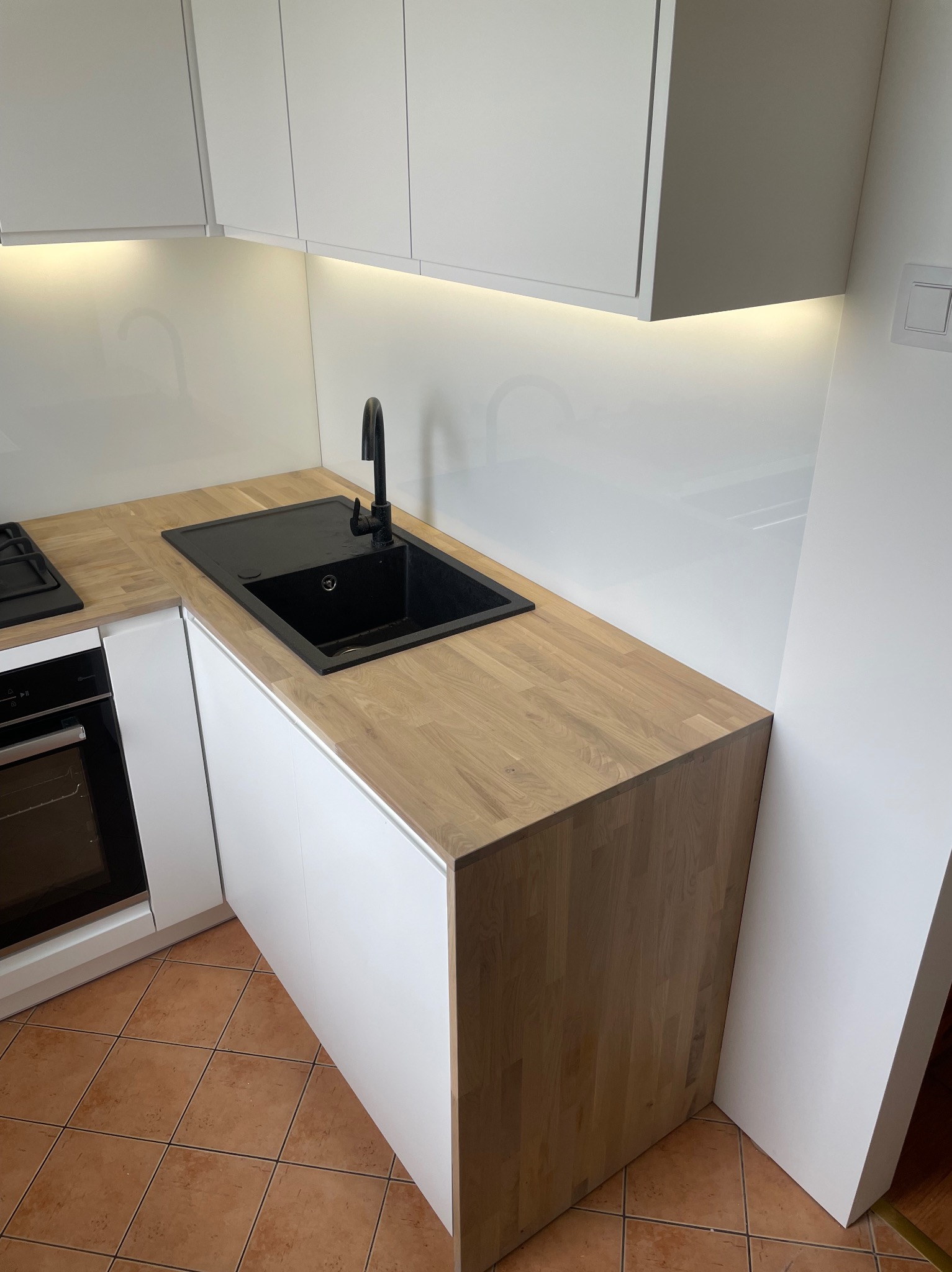 Nowoczesna kuchnia w Bujniczki-Kolonia: białe szafki z drewnianym blatem, czarny zlew i bateria, oświetlenie podszafkowe, brązowa terakota.