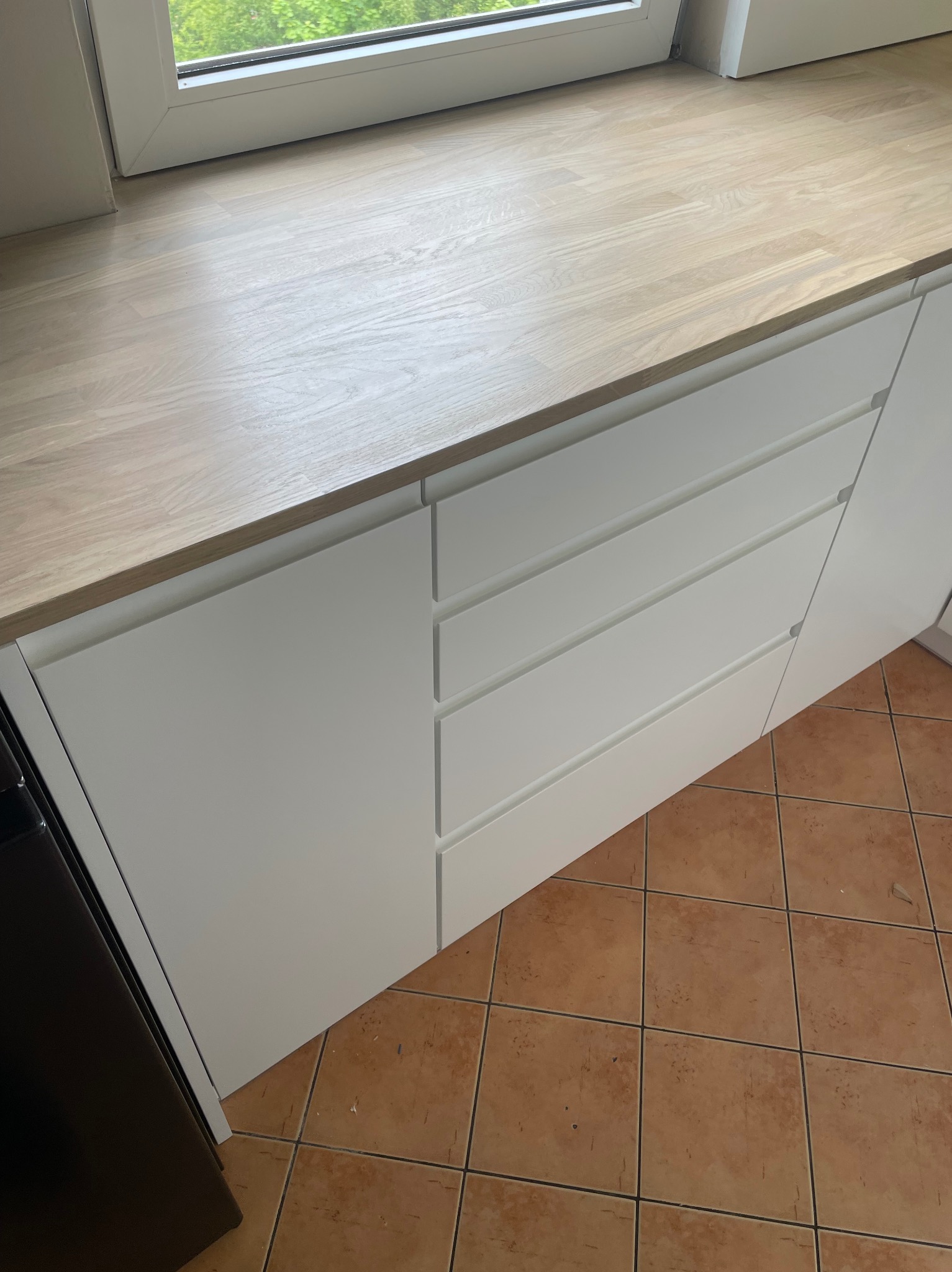 Blat z jasnego drewna przylegający do okna, pod nim białe szafki z trzema szufladami, na podłodze brązowe płytki.