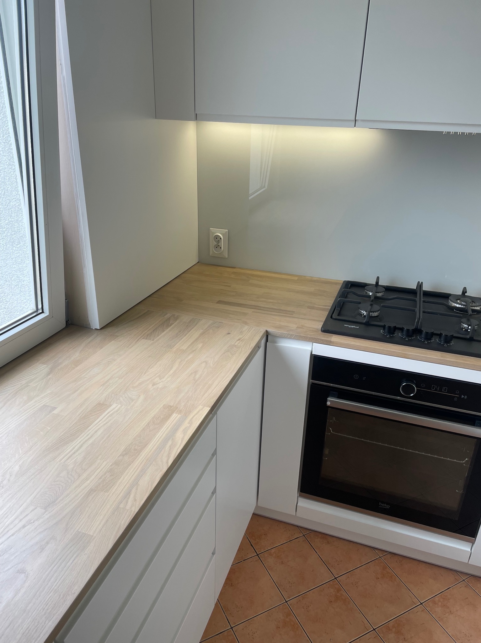 Fragment nowoczesnej kuchni z jasnym drewnianym blatem, białymi szafkami, piekarnikiem Beko i płytą gazową Whirlpool, oświetleniem podszafkowym i gniazdkiem elektrycznym na ścianie.