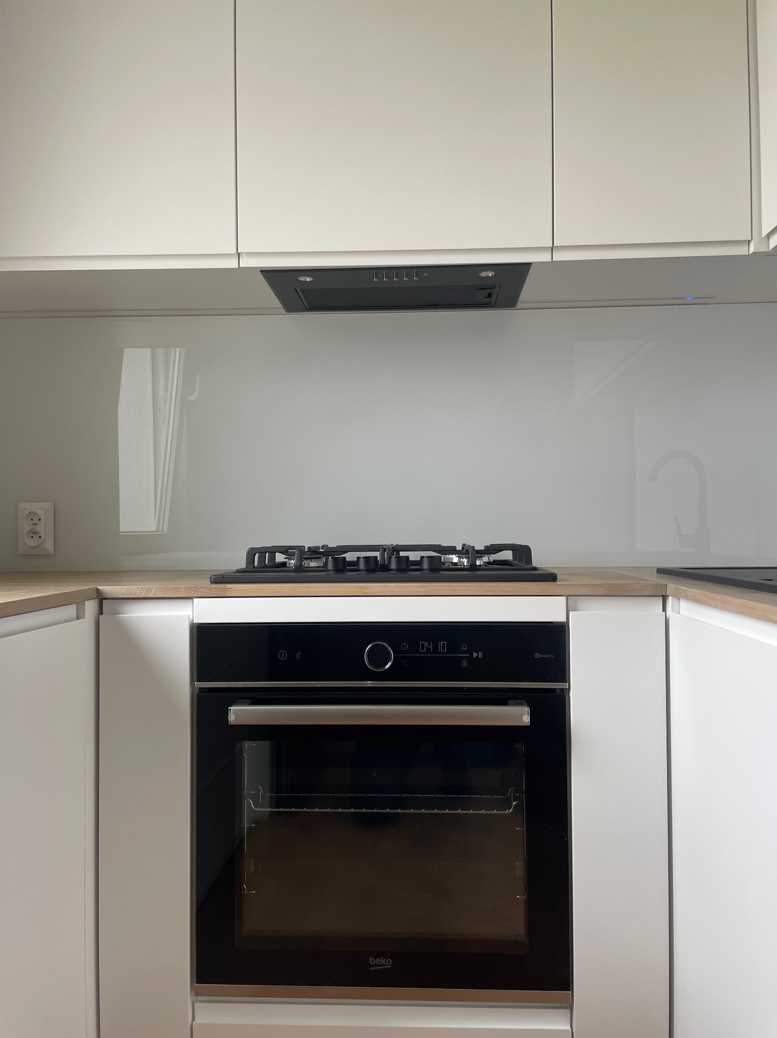Minimalistyczna kuchnia: białe szafki, drewniany blat, czarna płyta gazowa i piekarnik Beko z wyświetlaczem czasu, szklany panel nad blatem.