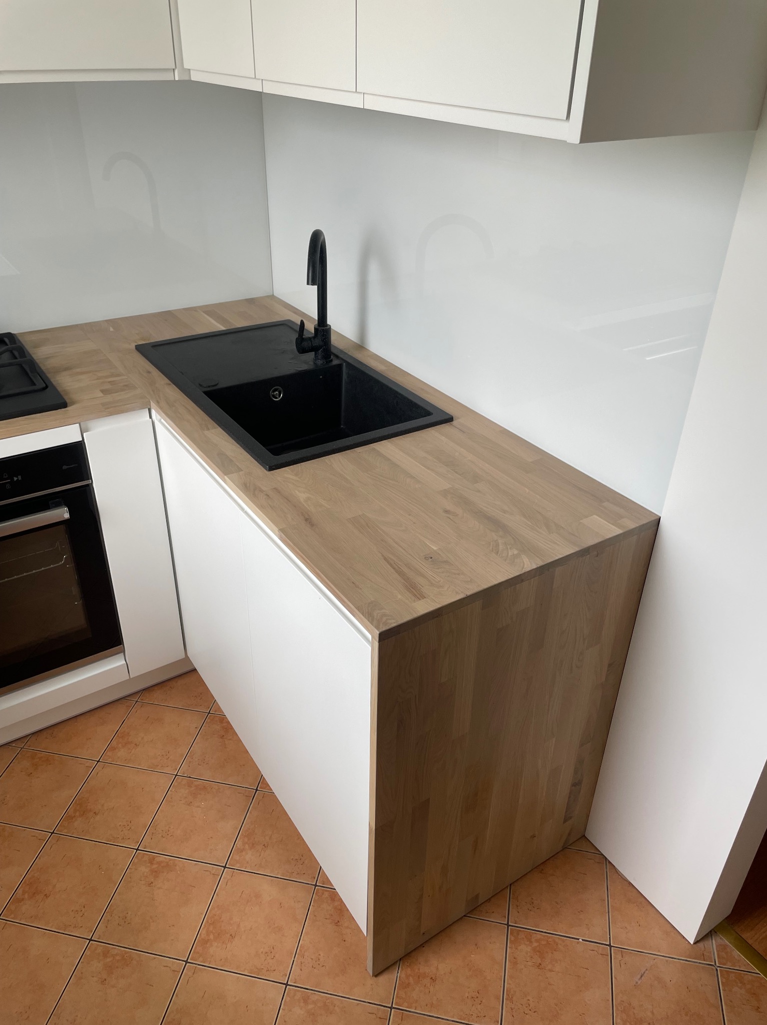Fragment nowoczesnej kuchni z drewnianym blatem, czarnym zlewozmywakiem i baterią, białe szafki, brązowa terakota na podłodze.