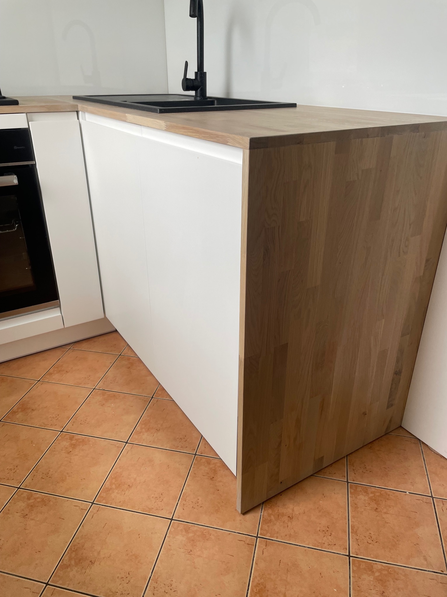 Fragment nowoczesnej zabudowy kuchennej z białymi frontami i blatem z jasnego drewna, czarny zlew i bateria, brązowa terakota na podłodze.