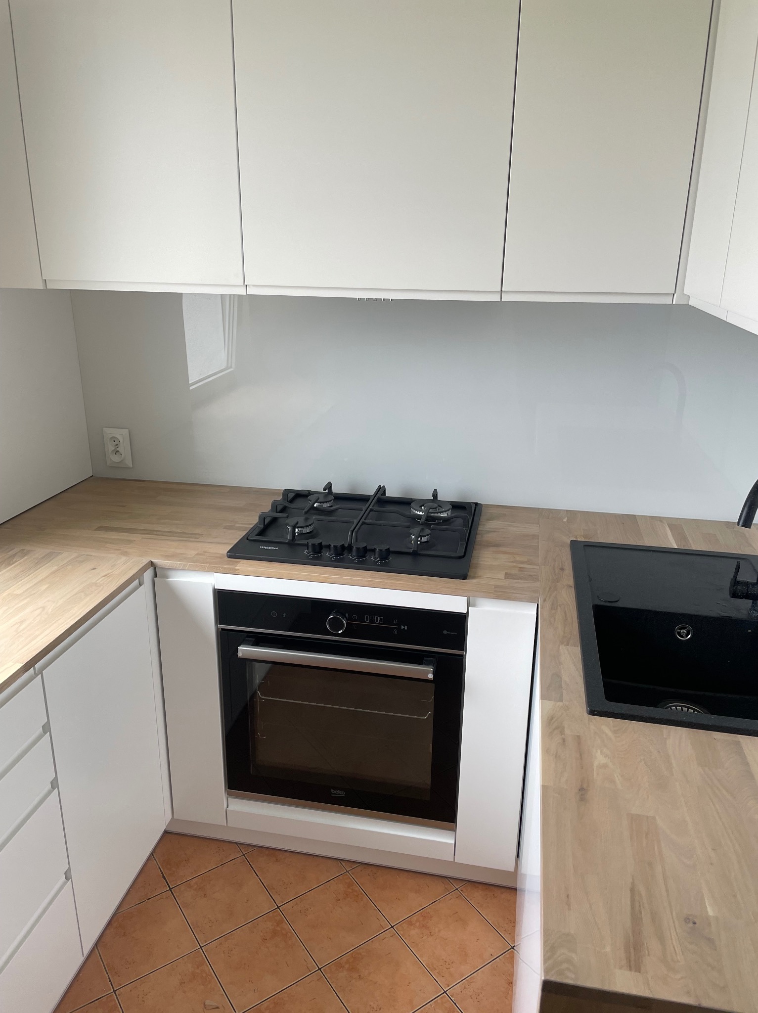 Nowoczesna kuchnia z białymi szafkami, drewnianym blatem, czarną płytą gazową i zlewem, oraz piekarnikiem marki Beko. Widoczny fragment brązowej podłogi z płytek.