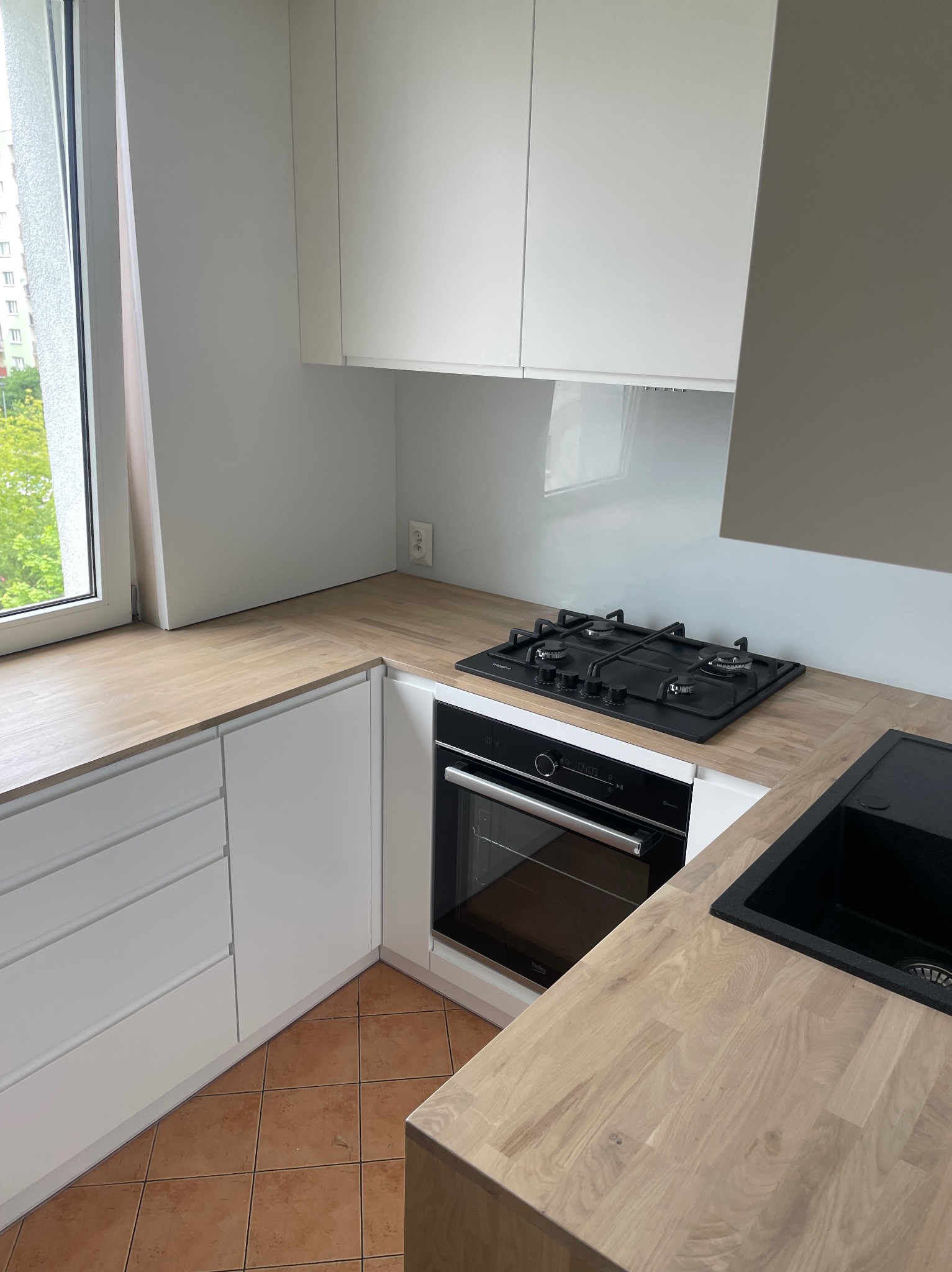 Nowoczesna kuchnia z białymi szafkami, drewnianym blatem, czarną płytą grzewczą i piekarnikiem; brązowa podłoga z terakoty, widok na zielone drzewa za oknem.