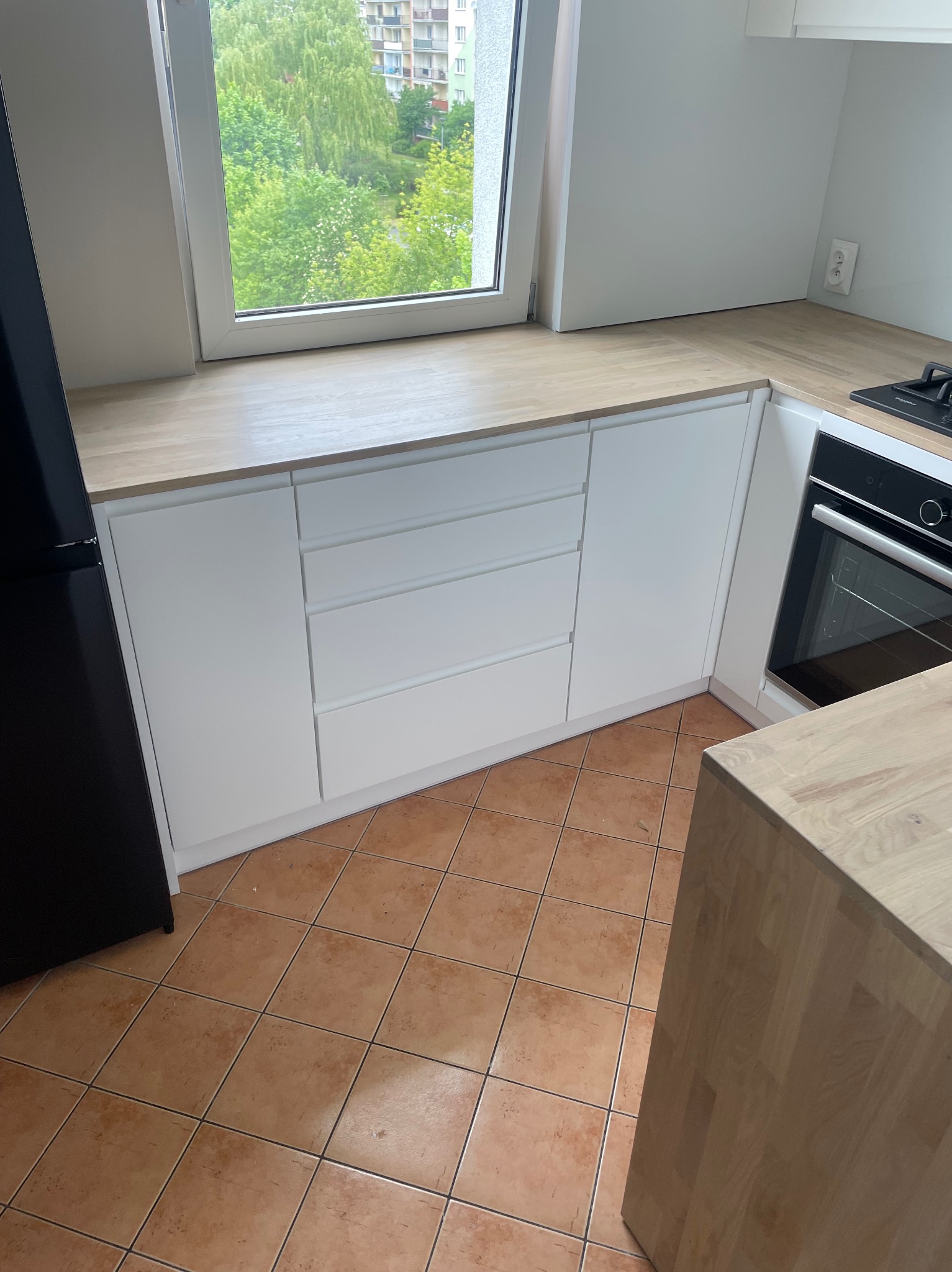 Nowoczesna, biała zabudowa kuchenna pod oknem z blatem w kolorze jasnego drewna, widok na zewnątrz przez okno, brązowa podłoga z płytek.