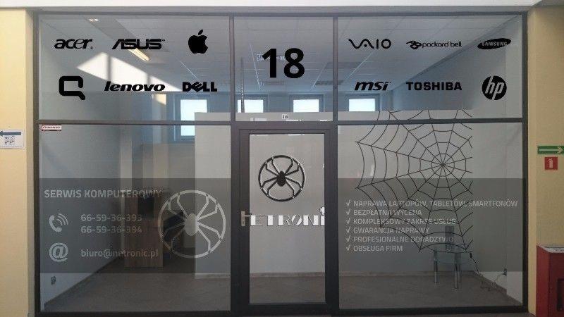 Szklane drzwi wejściowe do serwisu komputerowego z naklejonymi logotypami producentów laptopów, adres e-mail, numery telefonów, logo firmy z pająkiem oraz informacjami o naprawie laptopów, tabletów...