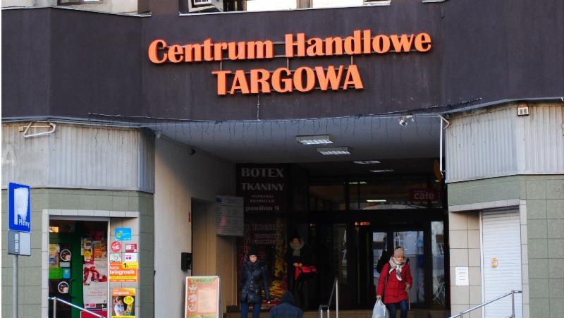 Wejście do Centrum Handlowego Targowa z neonowym napisem, widoczne osoby wchodzące i wychodzące, oraz tablice informacyjne i reklamy.