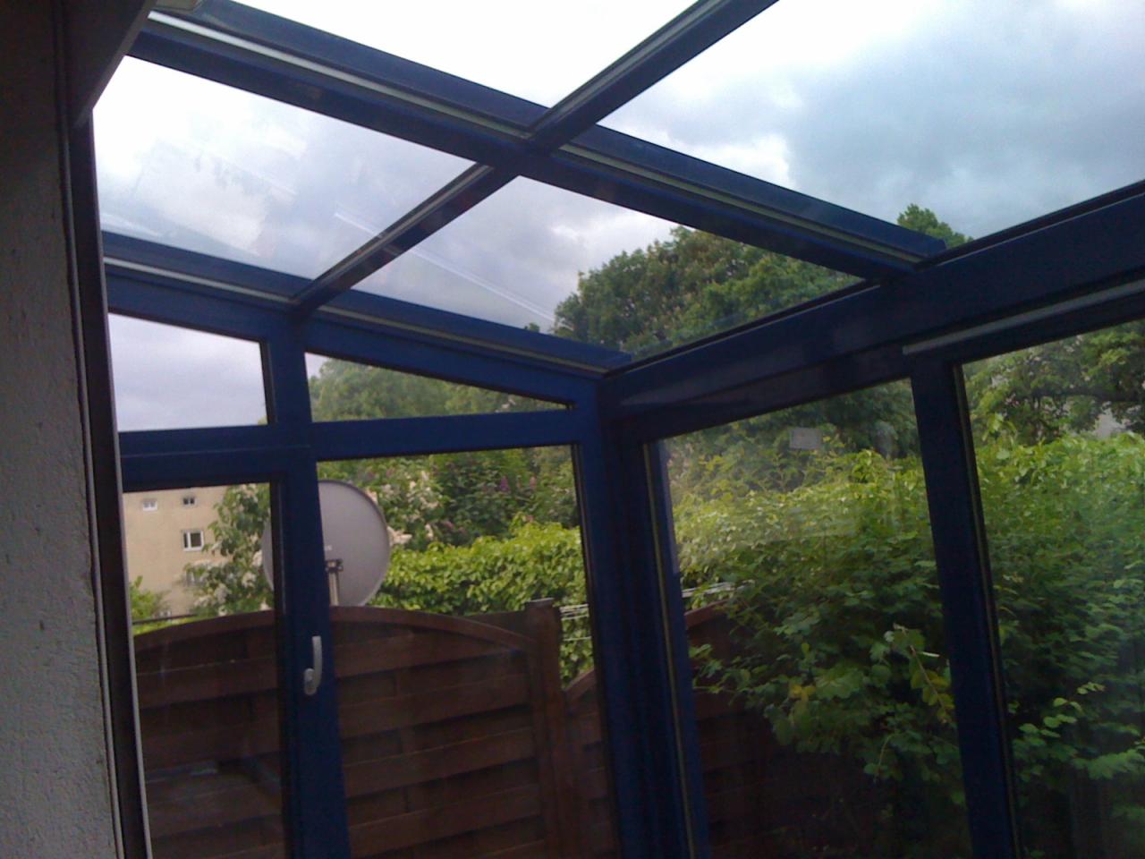 Zadaszenie tarasu z niebieskimi ramami okiennymi w Gdańsku, widok na ogród i pochmurne niebo.