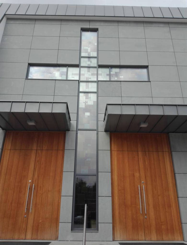 Nowoczesna fasada budynku z dużymi drewnianymi drzwiami wejściowymi i oknami w kształcie krzyża z dekoracyjnymi szybami.