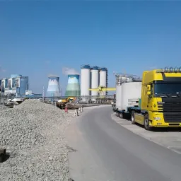 Żółty ciągnik siodłowy DAF z naczepą przewożący betonowe elementy na tle elektrowni z chłodniami kominowymi i hałdą kruszywa.