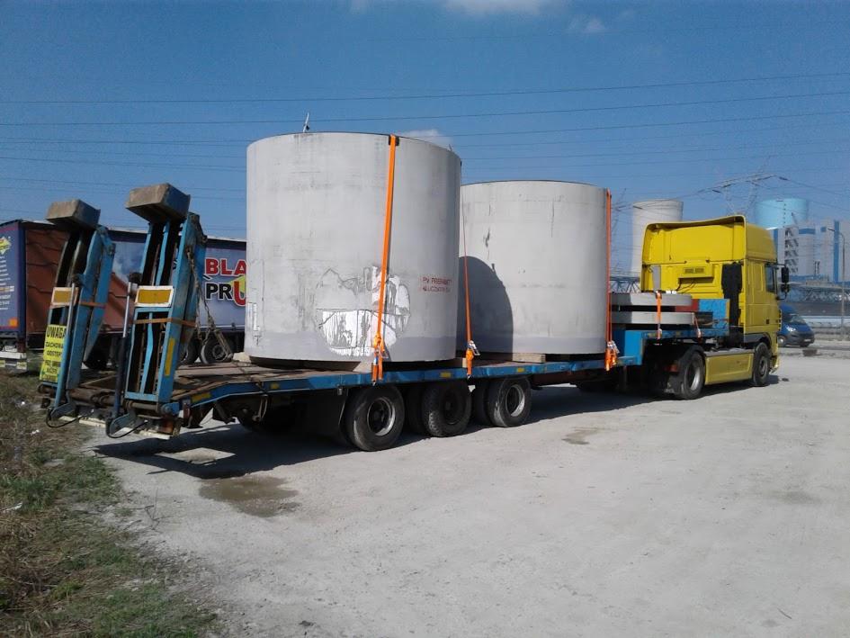 Żółta ciężarówka z naczepą przewożąca dwa duże, betonowe cylindry zabezpieczone pomarańczowymi pasami transportowymi na terenie przemysłowym w słoneczny dzień.