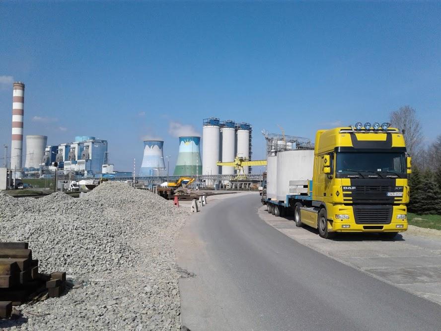 Żółty ciągnik siodłowy DAF z naczepą przewożący betonowe elementy na tle elektrowni z chłodniami kominowymi i hałdą kruszywa.