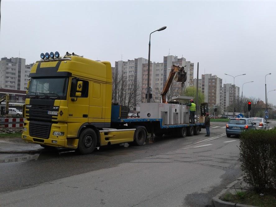Żółta ciężarówka DAF XF z dźwigiem ładuje betonowe elementy na naczepę na tle bloków mieszkalnych w pochmurny dzień.