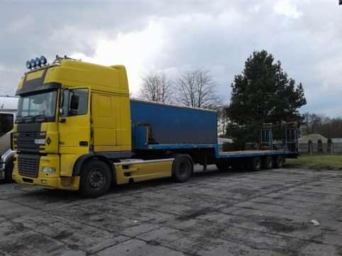 Żółta ciężarówka z niebieską naczepą na betonowym placu, widoczne drzewa w tle.