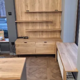 Drewniana zabudowa ściany z półkami i szafkami z szufladami, uzupełniona blatem roboczym przy oknie, na podłodze parkiet w jodełkę.