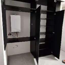 Czarna szafa na wymiar z otwartymi drzwiami, wnęka z piecem gazowym. Szare płytki na podłodze, białe ściany. Widoczne zawiasy i półki wewnątrz szafy.