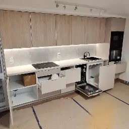 Jasna kuchnia z drewnopodobnymi szafkami i otwartymi szufladami, widoczne niedokończone wykończenie podłogi. Oświetlenie podszafkowe podkreśla blat.