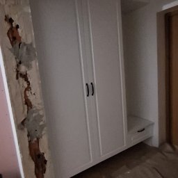 Mateusz Zdanowski - Biała szafa wnękowa z czarnymi uchwytami, widoczna na tle remontowanego pomieszczenia z odsłoniętą ścianą i drewnianą podłogą. Ujęcie z dołu.